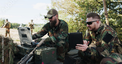 Soldiers in camouflage uniforms, equipped with helmets and backpacks, analyze battle strategy at the training ground, entering data into a system and discussing tactics while holding a tablet.