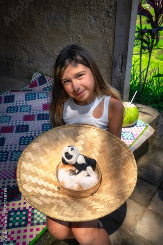 Obraz na płótnie A cheerful girl beams with joy while joyfully holding up a colorful decorative straw hat, featuring a fluffy panda toy and a kitten, all within a peaceful outdoor environment, Ubud, Bali, Indonesia w salonie