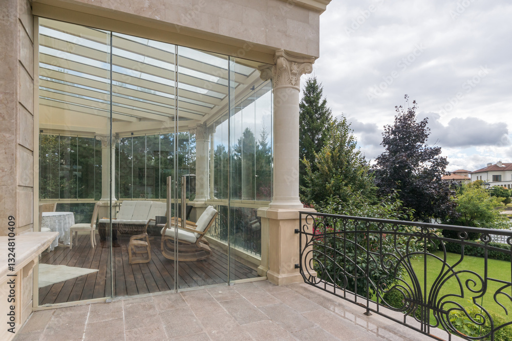 Fototapeta premium Glazed terrace of a luxurious country mansion decorated in neoclassical style. Stylish furniture. Beautiful view of the green lawn with trees.