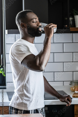Canvas Print An African American man in a white t-shirt and plaid pajama pants drinks a glass of water in a modern kitchen