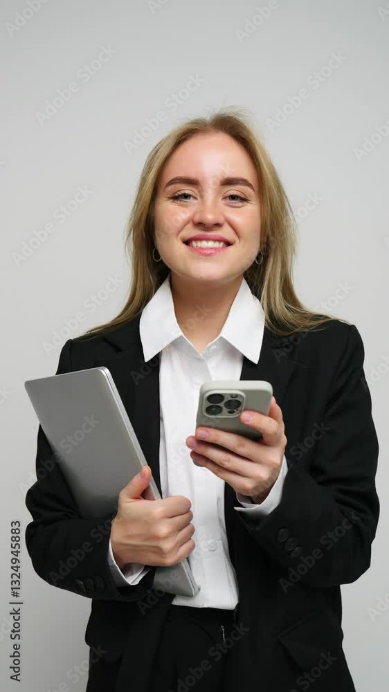 Smiling young woman in a blazer holds laptop and smartphone, ready for business tasks in a studio. Smiling young woman in a blazer holds laptop and smartphone, ready for business tasks in a studio.