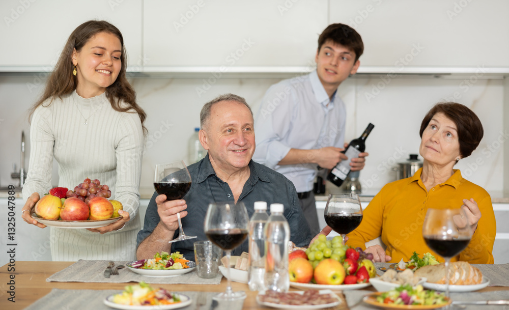 Happy carefree senior parents sharing memories with grown daughter and son-in-law during conversation over family celebration with wine at cozy home environment Happy carefree senior parents sharing memories with grown daughter and son-in-law during conversation over family celebration with wine at cozy home environment