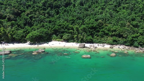 Aerial view of Ilha das Couves located in Ubatuba, Brazil. People enjoy the beach and the sea water crashes against the rocks on the coast. Green trees are seen in the beautiful landscape.
