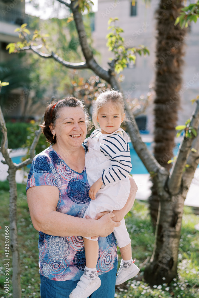 Smiling grandmother with a little girl in her arms stands near a tree in a green garden Smiling grandmother with a little girl in her arms stands near a tree in a green garden