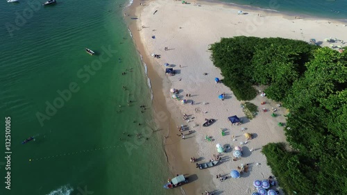 Aerial view of Ilha do Prumirim located in Ubatuba, Brazil. People enjoy the beach and the sea water crashes against the rocks on the coast. Green trees are seen in the beautiful landscape.