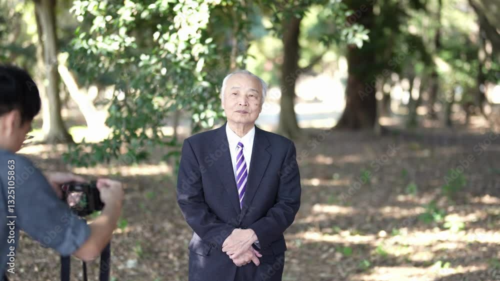 A man in his 30s is photographing a 78-year-old Japanese man in a suit standing and talking in a natural park filled with trees. A man in his 30s is photographing a 78-year-old Japanese man in a suit standing and talking in a natural park filled with trees.