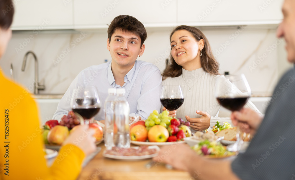 Cheerful young couple engaging in lively conversation with elderly parents at dinner table with wine and fruits in cozy home kitchen.. Cheerful young couple engaging in lively conversation with elderly parents at dinner table with wine and fruits in cozy home kitchen..