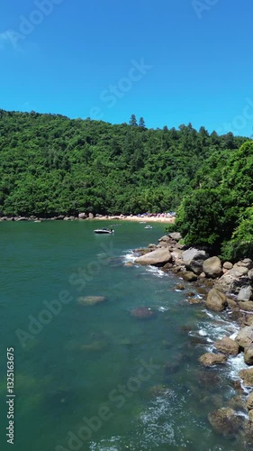 Aerial view of Praia do Cedro located in Ubatuba, Brazil. People enjoy the beach and the sea water crashes against the rocks on the coast. Green trees are seen in the beautiful landscape.