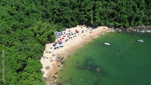 Aerial view of Praia do Cedro located in Ubatuba, Brazil. People enjoy the beach and the sea water crashes against the rocks on the coast. Green trees are seen in the beautiful landscape.