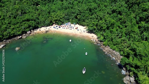 Aerial view of Praia do Cedro located in Ubatuba, Brazil. People enjoy the beach and the sea water crashes against the rocks on the coast. Green trees are seen in the beautiful landscape.