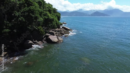 Aerial view of Praia do Cedro located in Ubatuba, Brazil. The sea water crashes against the rocks on the coast. Green trees are seen in the beautiful landscape.