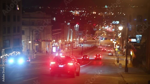 Cars stream through a brightly lit downtown street at night. Headlights and taillights create streaks of color. Buildings glow in the background. Smooth, steady shot.