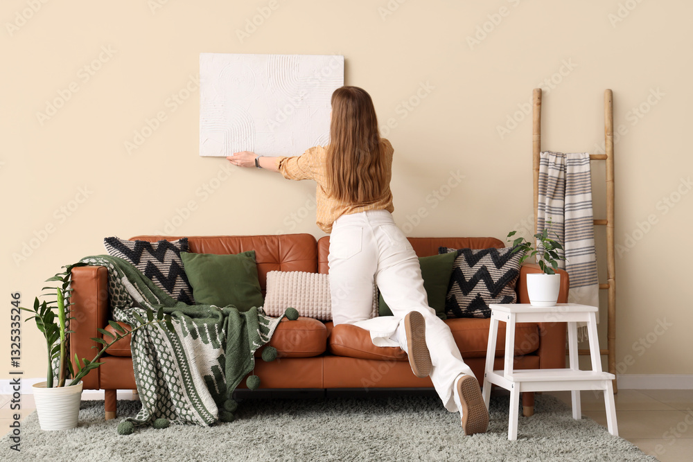 Fototapeta premium Young woman hanging picture in interior of living room. Back view