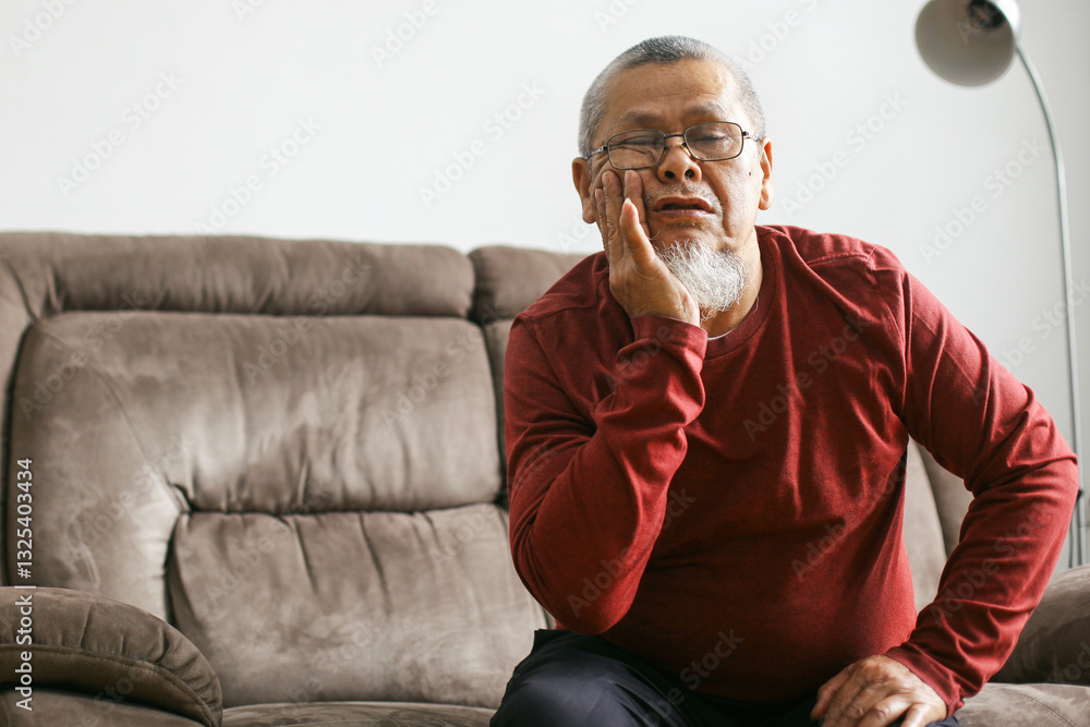 Elderly Asian Man Sits At Home, Touching His Cheek And Grimacing In Pain Suffering From Severe Toothache Or Jaw Discomfort Elderly Asian Man Sits At Home, Touching His Cheek And Grimacing In Pain Suffering From Severe Toothache Or Jaw Discomfort