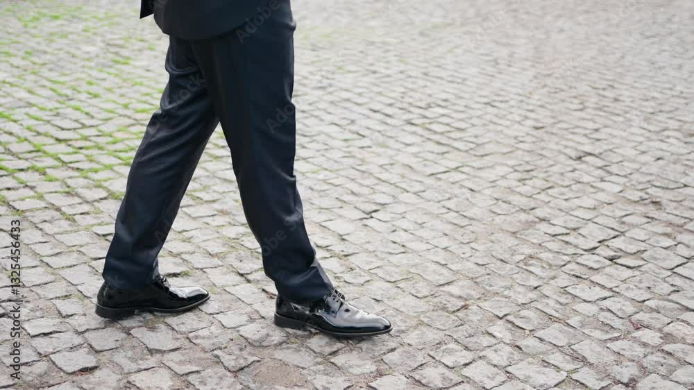 Man in black suit and polished shoes walking on cobblestone near a rustic stone wall Man in black suit and polished shoes walking on cobblestone near a rustic stone wall