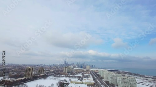 Aerial winter view of a sprawling cityscape with snow-covered trees and rooftops. High-rise buildings roads, parked cars, and distant neighborhoods stretch toward the horizon beneath a partly cloud