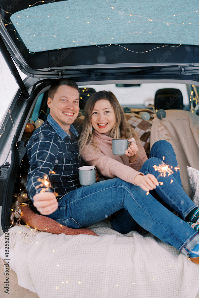 Smiling couple with mugs of coffee and sparklers in their hands are sitting on bedspreads in the trunk of a car