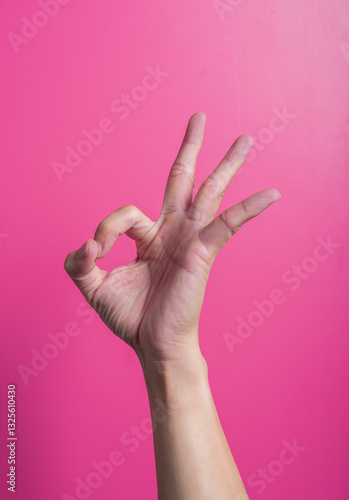 Asian hand making OK gesture with thumb and index forming a circle and three fingers raised, set against pink background, symbolizing approval, agreement, or positivity.