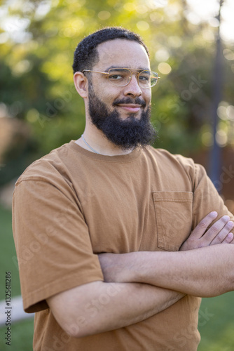 A cheerful man with glasses and a lovely beard stands confidently with his arms crossed, thoroughly enjoying a beautiful sunny day in a lush green outdoor environment surrounded by nature