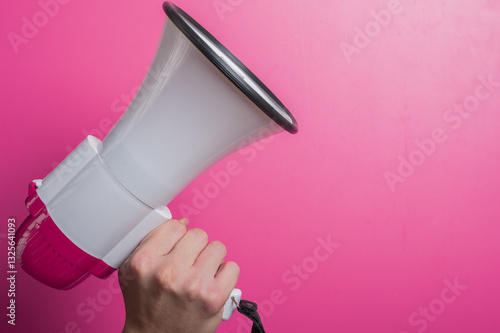 Asian hand holding a white and pink megaphone against a pink background, symbolizing speech, announcement, protest, or call to action in a minimalist studio setup.