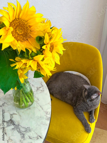 A grey cat is lying on a yellow armchair near a table with a bouquet of sunflowers