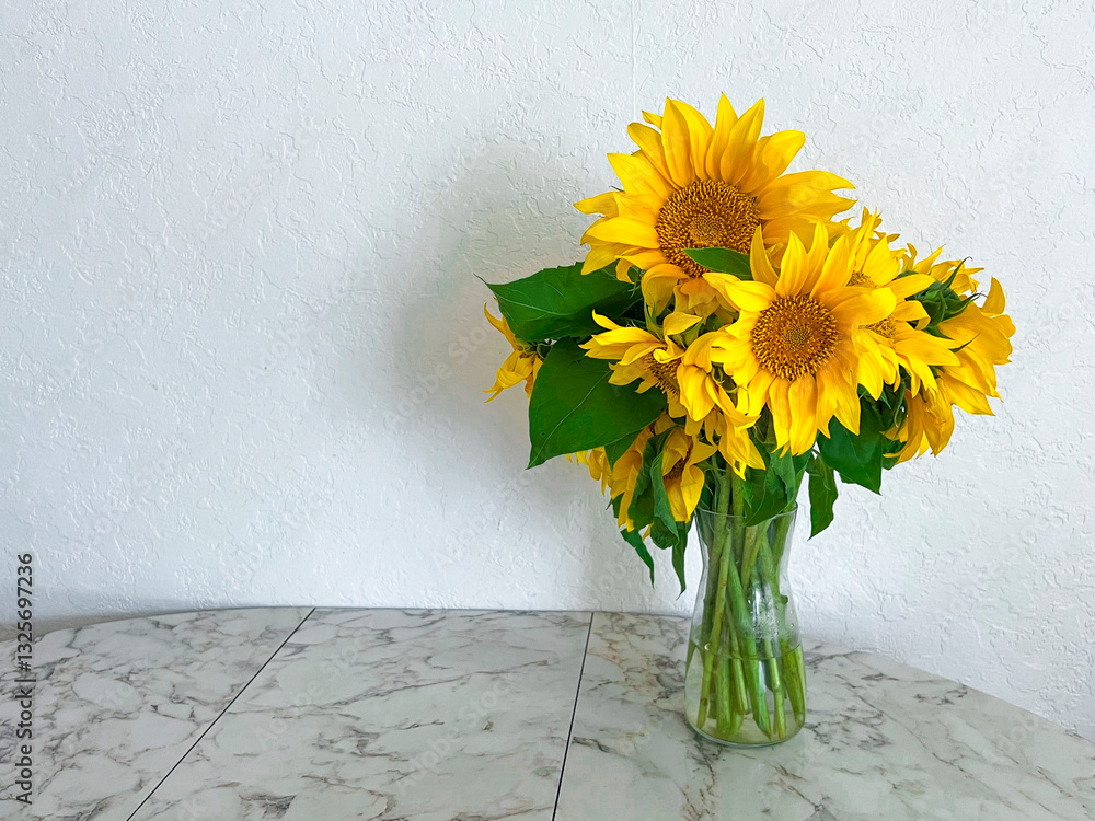 A bouquet of sunflowers stands in a glass vase on a marble table in a white kitchen