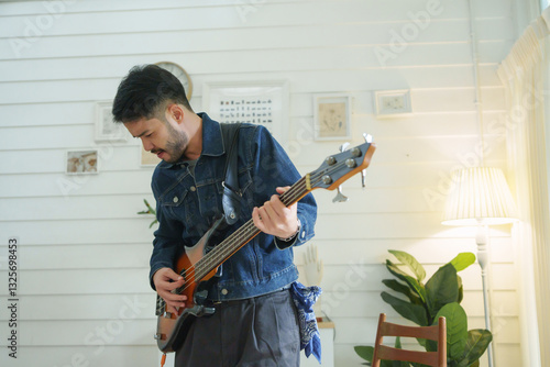 Принти на полотні Asian man in beard in denim jacket playing bass guitar passionately in living room, immersed in music with expressive posture and concentration