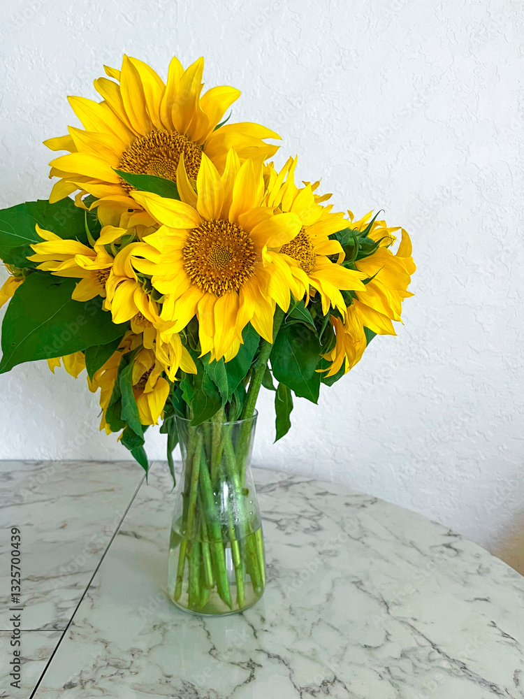 A bouquet of sunflowers stands in a glass vase on a marble table in a white kitchen