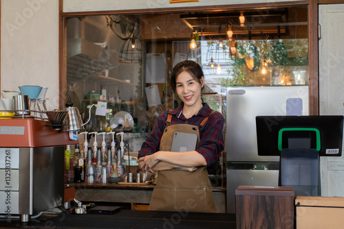 Asian beautiful businesswoman wears brown apron and holds tablet stands confidently behind counter of her newly opened café, entrepreneur owner coffee shop ready to serve customers.