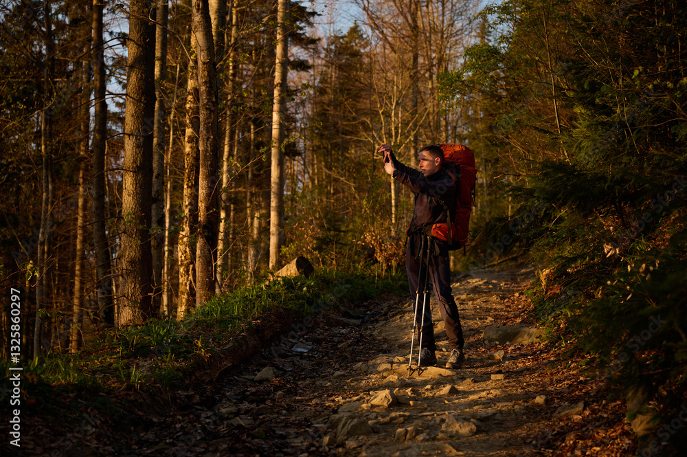 Fototapeta premium Fully equipped hiker taking pictures of sunrise on smartphone early in the morning in the mountains. Man standing with cell phone on mountain path enjoying sunrise.