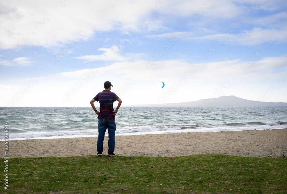 Man standing on Milford Beach. Unrecognizable people kiteboarding. Rangitoto Island in the distance. Auckland.