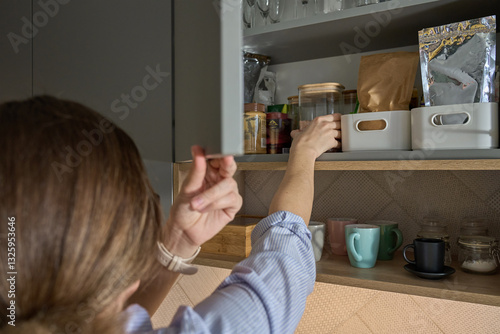 Obraz na płótnie Person takes jar from kitchen shelf filled with food containers and glassware. Woman open cupboard door in kitchen cabinet w salonie