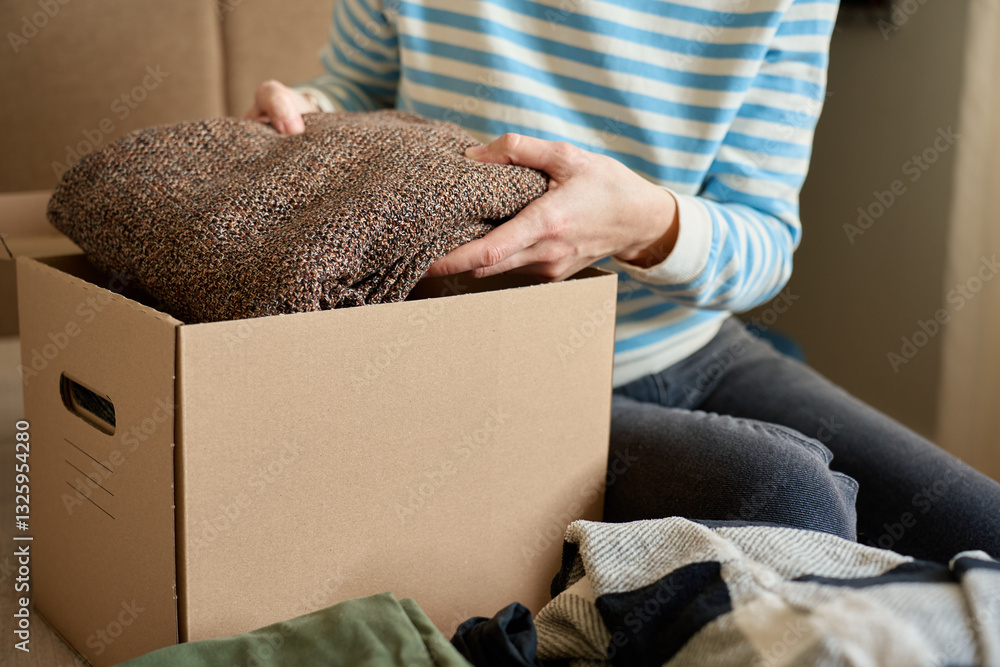 Woman packs donation box with clothes at home. Person places warm sweater into cardboard box. Preparing used clothing for recycling or second hand. Concept of charity and humanitarian aid