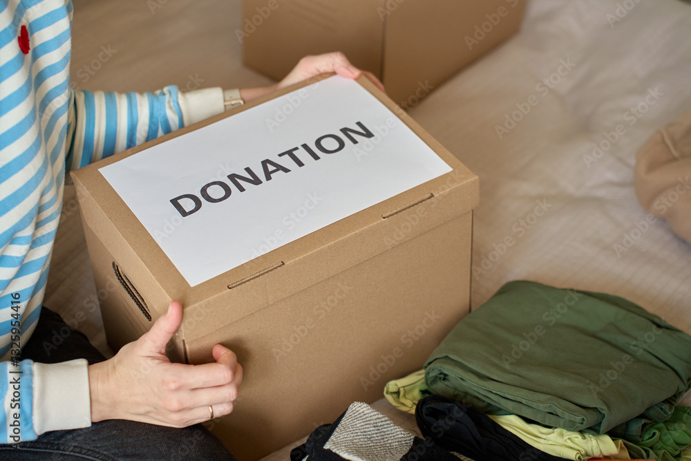 Woman holds donation box filled with clothes on bed, organizing items for charity. Cardboard box with clothes for recycling or second hand. Concept of volunteering, charity and social responsibility