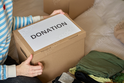 Obraz na płótnie Woman holds donation box filled with clothes on bed, organizing items for charity. Cardboard box with clothes for recycling or second hand. Concept of volunteering, charity and social responsibility w salonie
