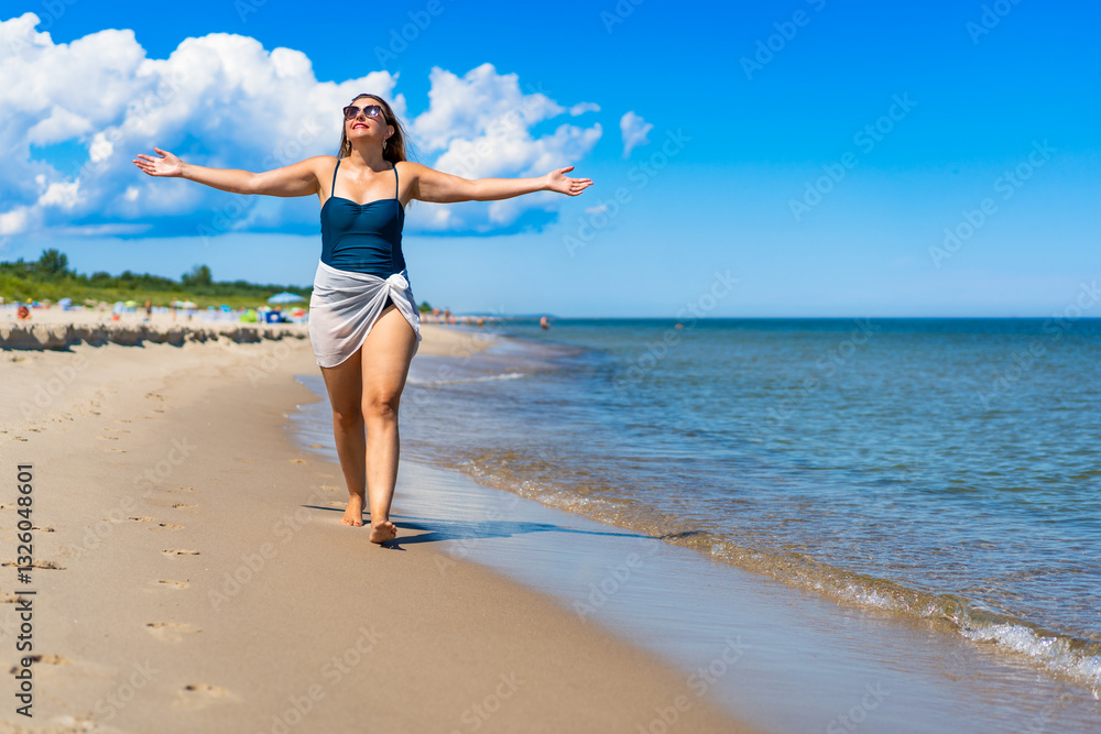 Beautiful middle-aged woman in swimsuit walking with outstretched arms on sandy beach in summertime. Energy and happiness. Front view Beautiful middle-aged woman in swimsuit walking with outstretched arms on sandy beach in summertime. Energy and happiness. Front view