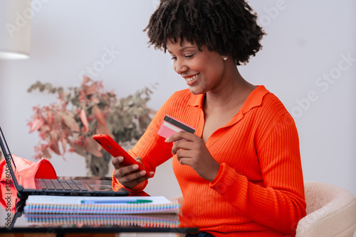 African american student making an online payment with a credit card while using a phone at home. Modern workspace with books, stationery, and stylish decor. E-commerce,digital transaction,and finance