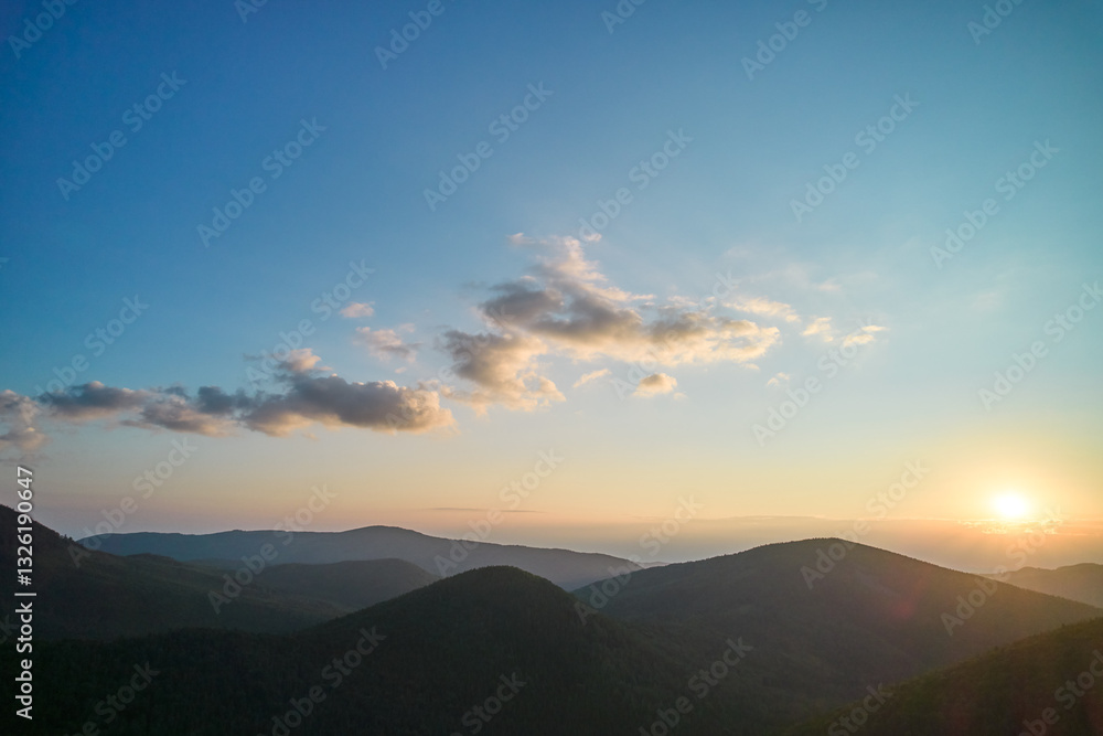 Obraz premium Sun sets behind rolling hills, casting warm, golden glow across sky. Wispy clouds, tinged with hues of orange and yellow, stretch across deepening blue sky. Serene and expansive landscape.