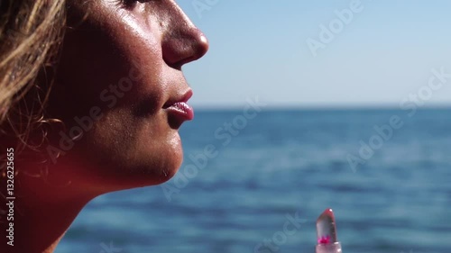 Woman, beach, lipstick - pretty lady applies protective lipstick for lip care. On beach, woman uses nourishing lipstick after day of sun exposure to refresh lips. Close up, Slow motion