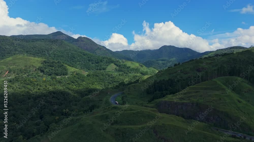 Aerial drone zoom in view of green mountain with vegetation, forest and trees with road where cars and trucks pass, blue sky with several white clouds in Brazil. Cloud shadow. Aerial drone zoom in view of green mountain with vegetation, forest and trees with road where cars and trucks pass, blue sky with several white clouds in Brazil. Cloud shadow.