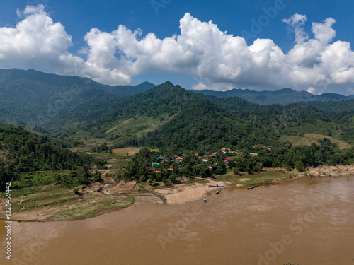 Aerial view of a picturesque rural landscape featuring a serene river, lush greenery, and a tranquil village surrounded by mountains and forests, Pak Tha District, Bokeo, Laos.