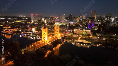Aerial view of illuminated tower bridge and vibrant cityscape at night with reflections on the river, The Bridge District, West Sacramento, California, United States.
