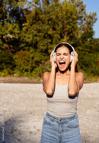 Young woman with headphones screaming loudly outdoors at nature alone. Releasing emotions and stress, silent scream practice, antistress technique
