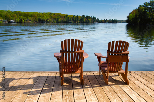 The calm blue lake in Muskoka ripples gently, likely from a passing boat. Cozy cabins sit among the trees, overlooking the water. Two Adirondack chairs rest on a wooden dock, facing the view.