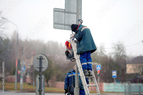 Obraz na płótnie Electrician standing on ladder repair traffic light. Worker fixing traffic light at busy intersection, using ladder and tools to perform maintenance. Road maintenance crew repairing traffic signal w salonie