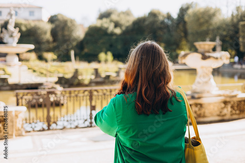 Back view of young female tourist near fountain, in Italy. Stylish woman visiting Italian landmarks. Traveling Europe, Italy in spring, rear view