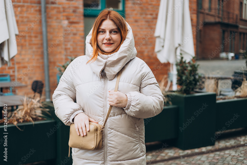 Stylish attractive young smiling 30s woman walking on city street in winter outfit, old town. Urban lifestyle concept. Portrait of happy woman looking at camera outdoor. High quality photo.