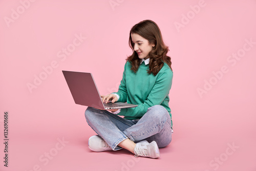  Young woman sitting on floor working on laptop. Smiling girl using laptop isolated on pink background.