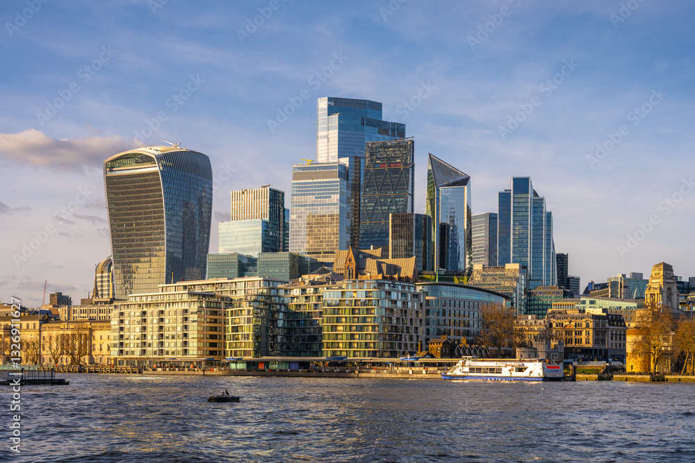 Obraz premium Modern skyscrapers rise against a clear sky in London City, reflecting on the River Thames. Boats glide along the water, showcasing the vibrant urban landscape of England.