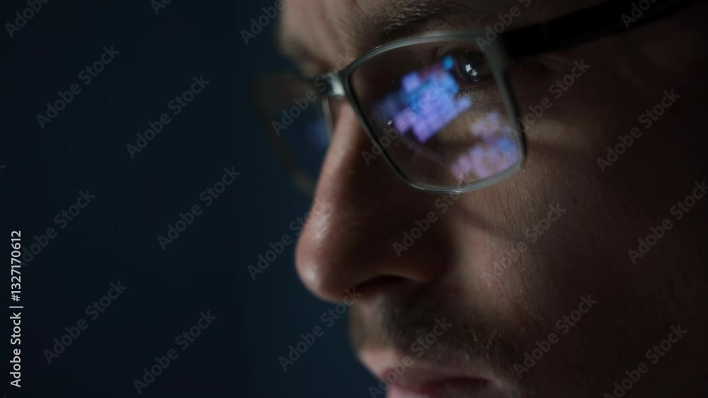 Close up of focused software engineer or data scientist wearing glasses, analyzing code on his screen. Programming script reflects on his eyeglasses as he works late.